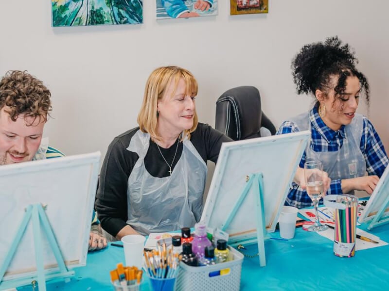 Three people smiling and painting at a paint and sip event